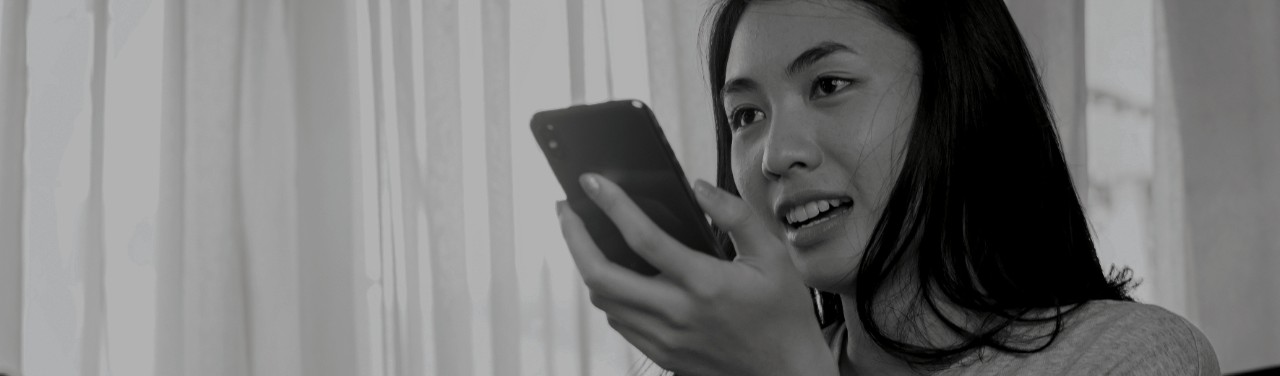 This photo shows a woman holding her mobile phone in her hand. The phone is held in front of her face, near her mouth. She is talking at the phone as if to ask the phone’s integrated virtual assistant a question. The photo suggests that online merchants must optimize for multilingual voice search if they want their products to be found on the crowded digital shelf. 